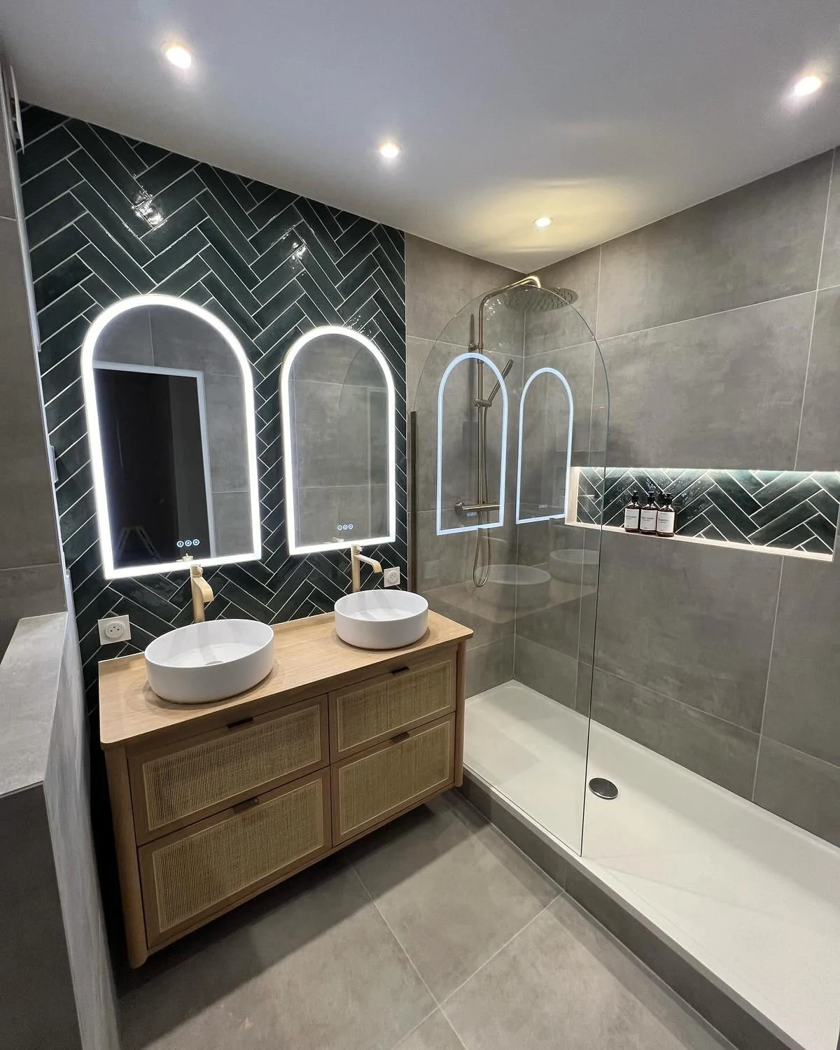 Salle de bain rénovée avec double vasque sur meuble en bois, miroirs LED en arche, carrelage vert posé en chevron et douche vitrée